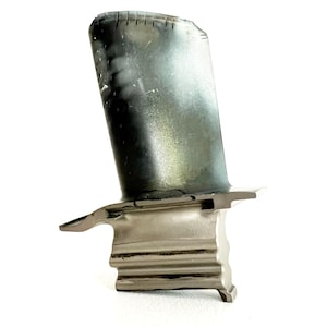 May include: A metallic gray turbine blade with a curved, elongated shape. The blade has a textured surface and a series of small notches along the top edge. The base of the blade is a lighter shade of gray with a ridged design.