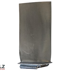 May include: A metallic grey turbine blade featuring a smooth, elongated rectangular body with a curved profile. The base of the component is wider and includes a distinct mechanical attachment point. The surface has a matte finish with subtle variations in tone and texture across the metal. The logo CLZ AVIATION is visible in the bottom left corner.