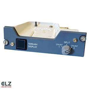 Pode incluir: Um painel de controle de aviação cinza com um botão quadrado preto rotulado TERRAIN DISPLAY e um botão giratório com configurações para LNAV 1, SPLIT e LNAV 2. A unidade possui uma carcaça de metal branco com parafusos visíveis e uma etiqueta.