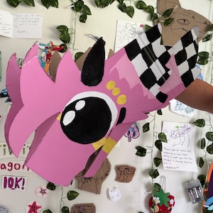 May include: A handcrafted cardboard dragon mask painted in bright pink with a large black eye, three yellow dots, and a black and white checkered pattern on the side. The mask features sharp, angular edges and a black horn-like protrusion on top.