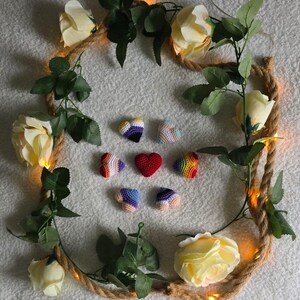 May include: A collection of crocheted hearts in various colors, including red, purple, blue, and rainbow hues, arranged on a white surface. The hearts are surrounded by a decorative wreath of rope, white roses, green leaves, and fairy lights.
