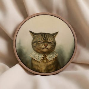 May include: A framed artwork features a grumpy-looking tabby cat wearing a lace collar with a pearl pendant. The cat's fur is brown and tan, and the artwork is set in a round wooden frame. The background is a soft, muted green.