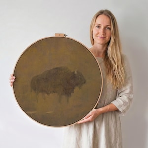 May include: A round, framed artwork featuring a bison in a sepia-toned landscape. The artwork is held by a person. The frame is made of light-colored wood. The bison is depicted in shades of brown and black.