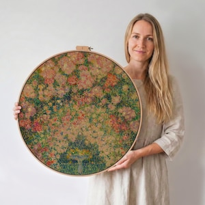 Könnte beinhalten: Ein runder floraler Stickrahmen mit einer farbenfrohen Gartenszene mit rosa, roten und grünen Blumen. Das Kunstwerk ist in einem natürlichen Holzrahmen gefasst, was eine rustikale und künstlerische Ästhetik erzeugt. Der Stickrahmen wird von einer Person gehalten.