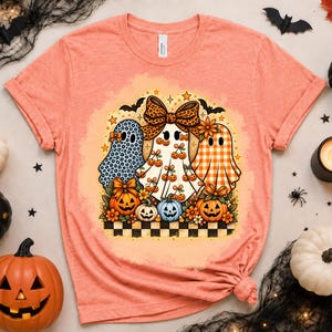 Peut inclure: Un t-shirt à manches courtes pêche chiné avec un motif central représentant trois fantômes. Les fantômes sont ornés d'imprimés léopard bleus, de motifs cerises et de vichy orange. Ils sont entourés de citrouilles, de fleurs et d'une base à damier.