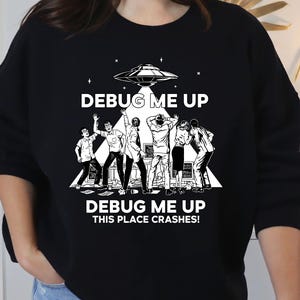 Peut inclure: Sweat-shirt noir avec un motif blanc représentant un OVNI et un groupe de personnes. Le texte "DEBUG ME UP" et "THIS PLACE CRASHES!" sont imprimés au-dessus et en dessous de l'illustration. Le design est une interprétation humoristique d'un thème technologique ou de science-fiction.