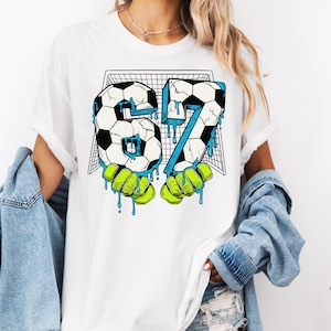 May include: White t-shirt featuring a soccer-themed graphic. The design includes the number "67" formed by soccer balls, with blue dripping accents and green goalie gloves. A soccer net is in the background.