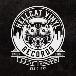 May include: A circular black and white logo featuring a stylized cat with vinyl record eyes. The text reads HELLCAT VINYL RECORDS, UNION STREET, SEATTLE WASHINGTON, and EST'D 1977. The design includes a city skyline silhouette and bridge elements.