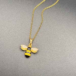 May include: Gold-toned necklace featuring a bee pendant. The bee pendant has a black and yellow striped body, gold wings with clear crystal accents, and a gold-toned chain. The necklace is displayed against a dark gray background.