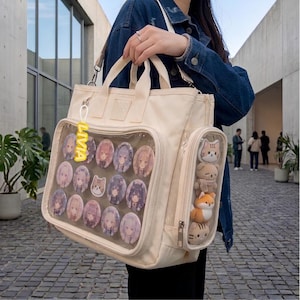 May include: A cream-colored canvas tote bag featuring a large clear front display pocket filled with circular anime-style character pins and a small cat sticker. A vertical side pocket contains four small plush animal toys and a yellow LIVIA keychain.