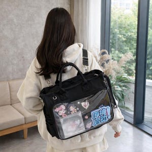 Könnte beinhalten: Schwarze Tragetasche mit einer transparenten Vordertasche, die Anime-Charakterkunst und japanischen Text zeigt. Die Tasche hat einen oberen Griff und verstellbare Schultergurte.