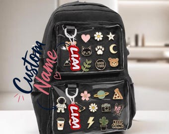 Puede incluir: Mochila negra con bolsillos transparentes llenos de pines coloridos y un llavero rojo que dice "LIAM". La mochila tiene las palabras "Custom Name" escritas en el lateral.
