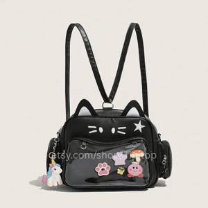 Puede incluir: Mochila negra con forma de gato con detalles blancos, incluyendo orejas y una estrella. Cuenta con un bolsillo frontal transparente con amuletos de dibujos animados coloridos y un llavero de unicornio. La mochila tiene correas ajustables y bolsillos laterales.
