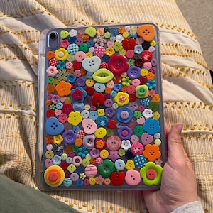 Puede incluir: Una funda transparente para iPad decorada con una variedad de botones coloridos. Los botones tienen varias formas y tamaños, incluyendo flores, corazones y círculos. La funda está sujeta en la mano de una persona.