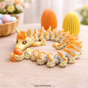 May include: A whimsical, cream-colored dragon figurine with orange accents and floral details. The dragon is coiled, with a horse-like head and a segmented body. Two decorative, textured egg-shaped objects are in the background. The text "CLYR 3D STUDIO" is at the bottom.