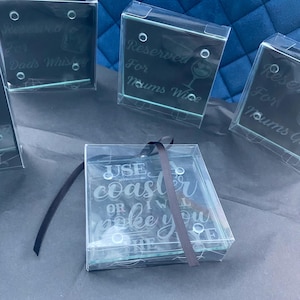 Op de afbeelding: Transparante glazen onderzetters met geëtste tekst, verpakt in transparante dozen. Eén onderzetter luidt "Use coaster or I will poke you". Andere zeggen "Reserved for Dads Whisky" en "Reserved for Mums Wine".