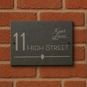 May include: A rectangular dark grey slate house sign featuring the engraved number 11, the text Kent Loves..., and the text HIGH STREET. The sign includes a small diamond shape above a horizontal line and is secured with two black screws.