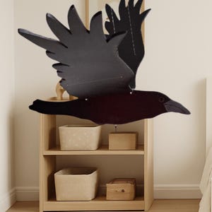 May include: A black raven mobile with outstretched wings, suspended in mid-air. The bird is made of dark material, with detailed wing and feather cutouts. The background includes a light wooden shelf with storage baskets and a framed picture.