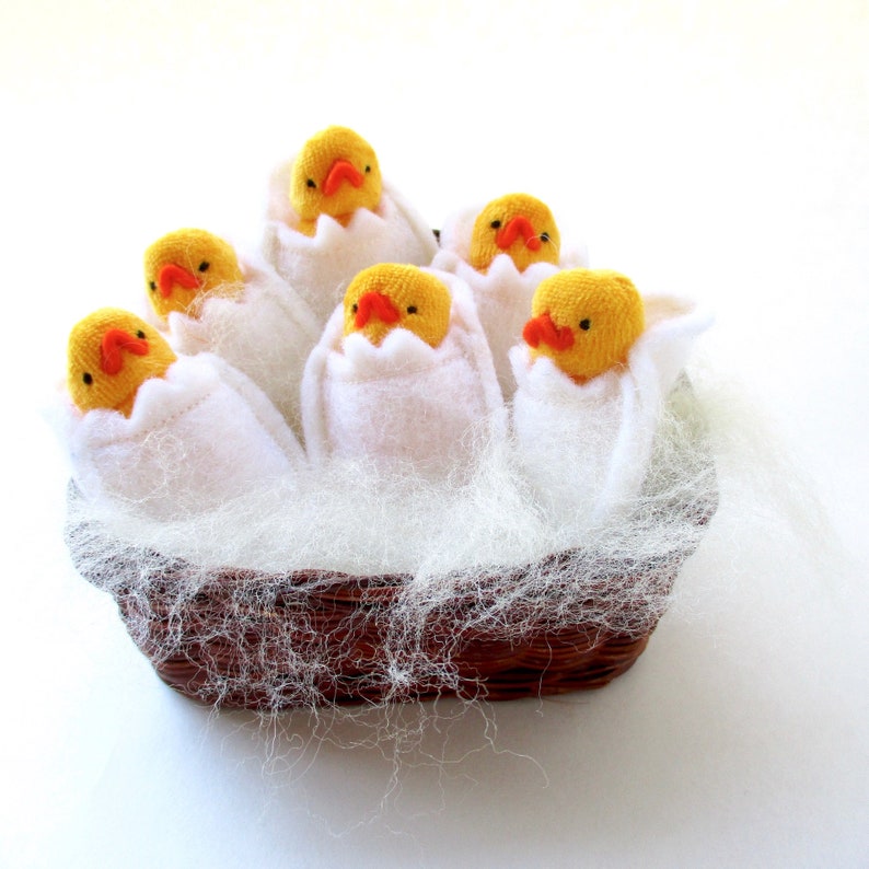 Op de afbeelding: Een decoratieve mand gevuld met zes gele kuikens die uit witte vilten eierschalen komen. De kuikens hebben oranje snavels en nestelen in een bed van witte vezels. De mand is bruin en heeft een geweven textuur.