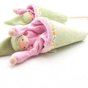 May include: Two plush dolls in green and pink cone-shaped holders. The dolls have round faces with small features and are wearing matching pink outfits. The holders are trimmed with a floral ribbon.