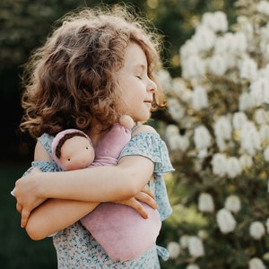 Pode incluir: Uma menina com cabelo castanho encaracolado abra&ccedil;a uma boneca macia cor de lavanda. A boneca tem um rosto simples e est&aacute; embrulhada numa manta de lavanda a condizer. A menina usa um vestido azul floral, criando uma cena serena e suave.