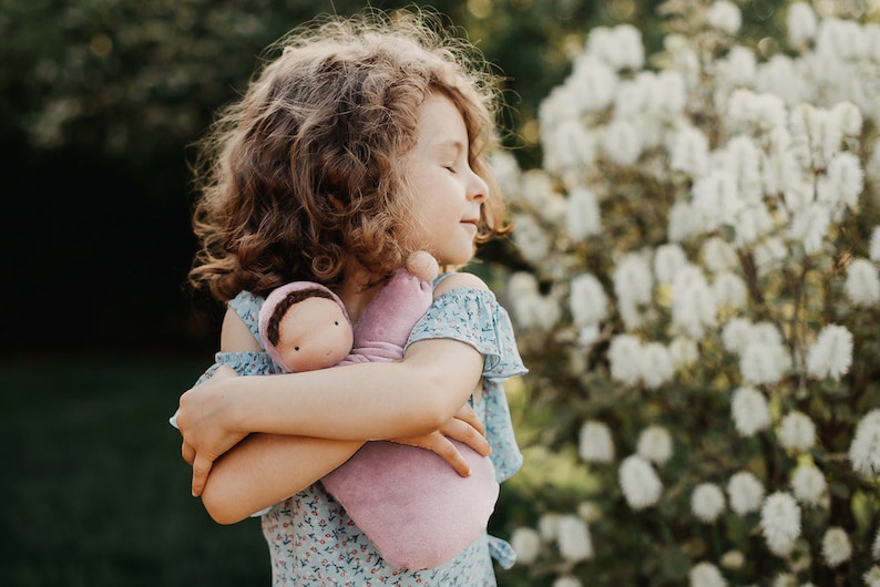 Pode incluir: Uma menina com cabelo castanho encaracolado abra&ccedil;a uma boneca macia cor de lavanda. A boneca tem um rosto simples e est&aacute; embrulhada numa manta de lavanda a condizer. A menina usa um vestido azul floral, criando uma cena serena e suave.