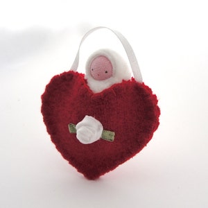 May include: A handmade red heart-shaped ornament with a white rose and a small doll figure. The doll has a pink face and is wearing a white hooded outfit. A white ribbon is attached for hanging.