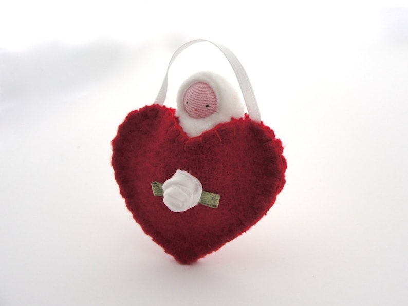 May include: A handmade red heart-shaped ornament with a white rose and a small doll figure. The doll has a pink face and is wearing a white hooded outfit. A white ribbon is attached for hanging.