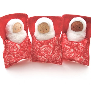 May include: Three handmade dolls, each nestled in a red fabric bed with a floral pattern. The dolls have white knitted swaddling and tan or brown faces with simple stitched features. The beds are a vibrant red.