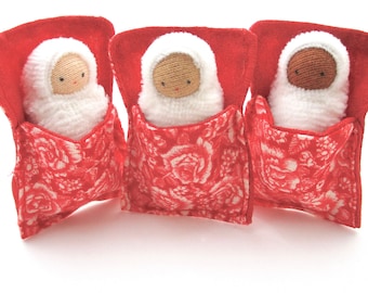 May include: Three handmade dolls, each nestled in a red fabric bed with a floral pattern. The dolls have white knitted swaddling and tan or brown faces with simple stitched features. The beds are a vibrant red.