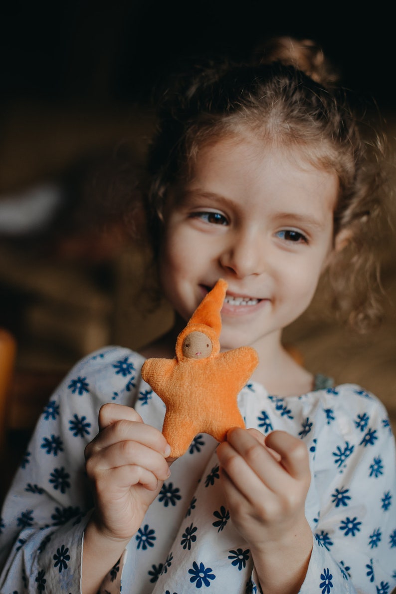 May include: A young girl with curly brown hair smiles while holding a small orange felt star-shaped doll with a pointed hat.