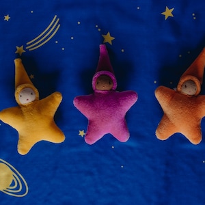 May include: Three plush star-shaped toys with embroidered faces and pointy hats. The stars are yellow, pink, and orange, and are lying on a blue fabric background with yellow stars.