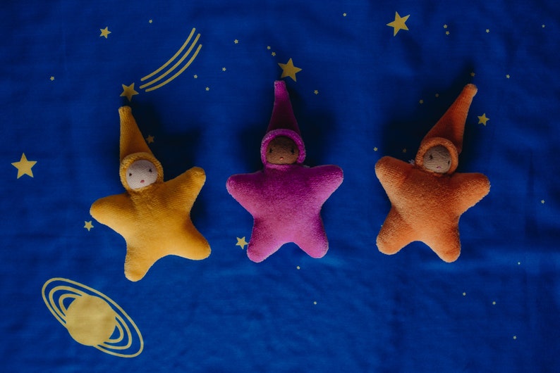May include: Three plush star-shaped toys with embroidered faces and pointy hats. The stars are yellow, pink, and orange, and are lying on a blue fabric background with yellow stars.
