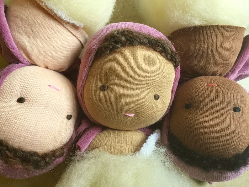 Pode incluir: Close-up de v&aacute;rias cabe&ccedil;as de bonecas feitas &agrave; m&atilde;o. As bonecas t&ecirc;m olhos castanhos, l&aacute;bios rosados e cabelo castanho. Algumas bonecas usam capuzes rosa. As bonecas s&atilde;o feitas de tecido e est&atilde;o em v&aacute;rias fases de conclus&atilde;o.