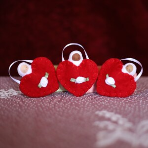 May include: Three red felt heart ornaments with white ribbon hangers. Each heart features a small doll figure and a white rose with green leaves. The ornaments are displayed on a patterned surface.