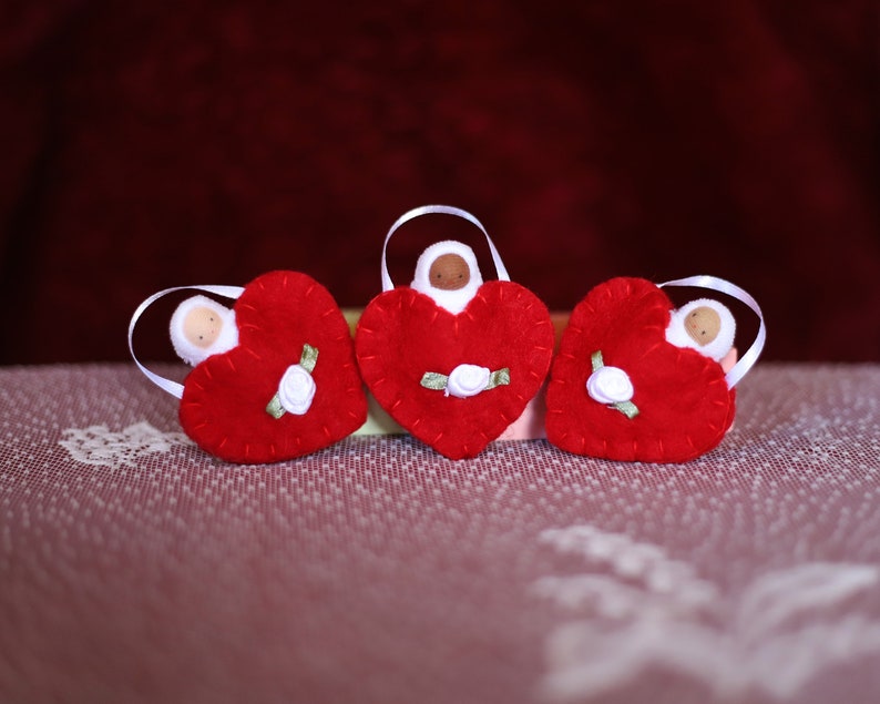 May include: Three red felt heart ornaments with white ribbon hangers. Each heart features a small doll figure and a white rose with green leaves. The ornaments are displayed on a patterned surface.