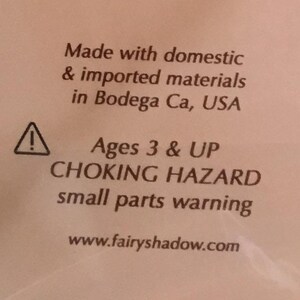 Op de afbeelding: Tekst op een witte achtergrond: "Made with domestic & imported materials in Bodega Ca, USA." Hieronder een waarschuwingssymbool en de tekst: "Ages 3 & UP CHOKING HAZARD small parts warning." De website www.fairyshadow.com staat ook vermeld.