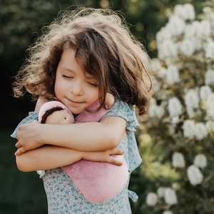 Pode incluir: Uma menina com cabelo castanho encaracolado abra&ccedil;a uma boneca rosa macia. A boneca est&aacute; embrulhada numa manta rosa a condizer e usa um pequeno gorro com cabelo castanho. A menina usa um vestido azul floral, com uma express&atilde;o serena.