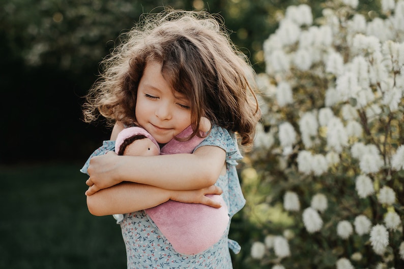 Pode incluir: Uma menina com cabelo castanho encaracolado abra&ccedil;a uma boneca rosa macia. A boneca est&aacute; embrulhada numa manta rosa a condizer e usa um pequeno gorro com cabelo castanho. A menina usa um vestido azul floral, com uma express&atilde;o serena.