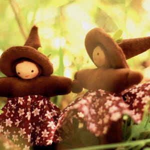 May include: Two handmade dolls with brown hats and arms, and red skirts with white floral patterns. The dolls have painted faces and are set against a blurred green background, creating a whimsical, fairytale-like scene.