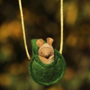May include: A small, brown, felt mouse with a green felt pouch hanging from a gold cord. The mouse is sitting inside the pouch.