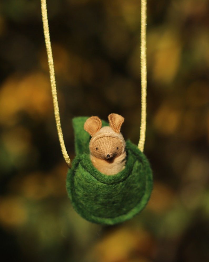 May include: A small, brown, felt mouse with a green felt pouch hanging from a gold cord. The mouse is sitting inside the pouch.