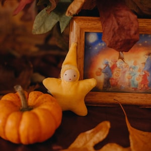 May include: A small yellow star-shaped doll with a white face and a pumpkin on a brown surface with dried leaves.