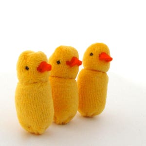 Op de afbeelding: Drie handgemaakte gele kuikenfiguren met oranje snavels en zwarte ogen. De kuikens zijn gemaakt van een zacht, getextureerd materiaal, mogelijk stof of garen. Ze staan rechtop tegen een witte achtergrond.