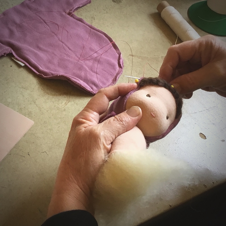 Pode incluir: Cria&ccedil;&atilde;o de uma boneca artesanal. O corpo e a cabe&ccedil;a s&atilde;o roxos claros. O rosto &eacute; bege claro, com cabelo castanho e olhos pretos pequenos. Enchimento branco e materiais de costura s&atilde;o vis&iacute;veis.