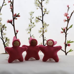 May include: Three pink felt star-shaped dolls with embroidered faces and pointy hats. The dolls are arranged in a row on a white surface in front of a background of white flowering branches.