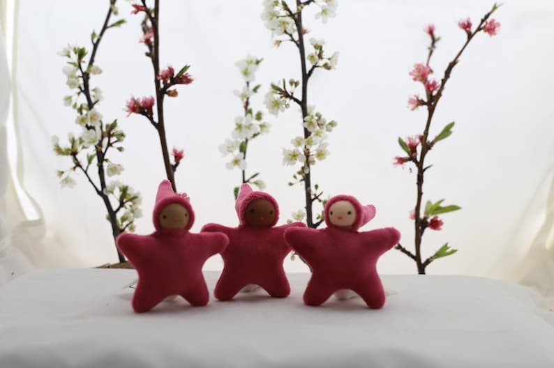 May include: Three pink felt star-shaped dolls with embroidered faces and pointy hats. The dolls are arranged in a row on a white surface in front of a background of white flowering branches.