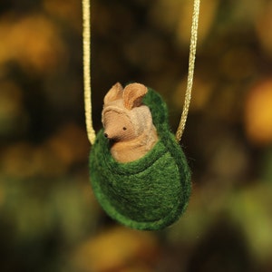 May include: A small, brown, felt animal with large ears is nestled inside a green felt pouch. The pouch is attached to a gold chain.