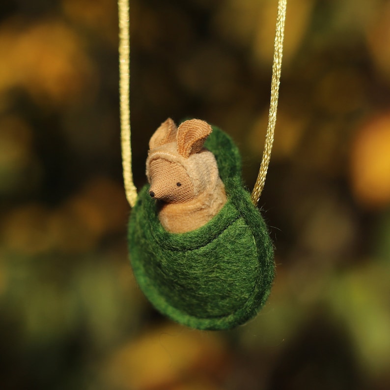 May include: A small, brown, felt animal with large ears is nestled inside a green felt pouch. The pouch is attached to a gold chain.