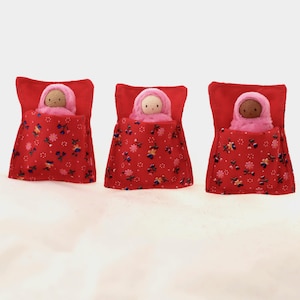 May include: Three handmade dolls in miniature beds. Each doll is nestled in a red bed with a matching pillow and floral patterned blanket. The dolls have pink hoods and are in a sleeping position.